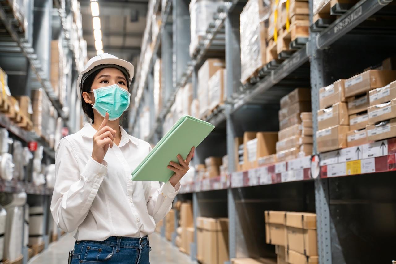 Empleada de almacén con mascarilla y tableta, realizando inspecciones de stock, ilustrando el enfoque de Del Bravo en la precisión y seguridad en la gestión de inventarios